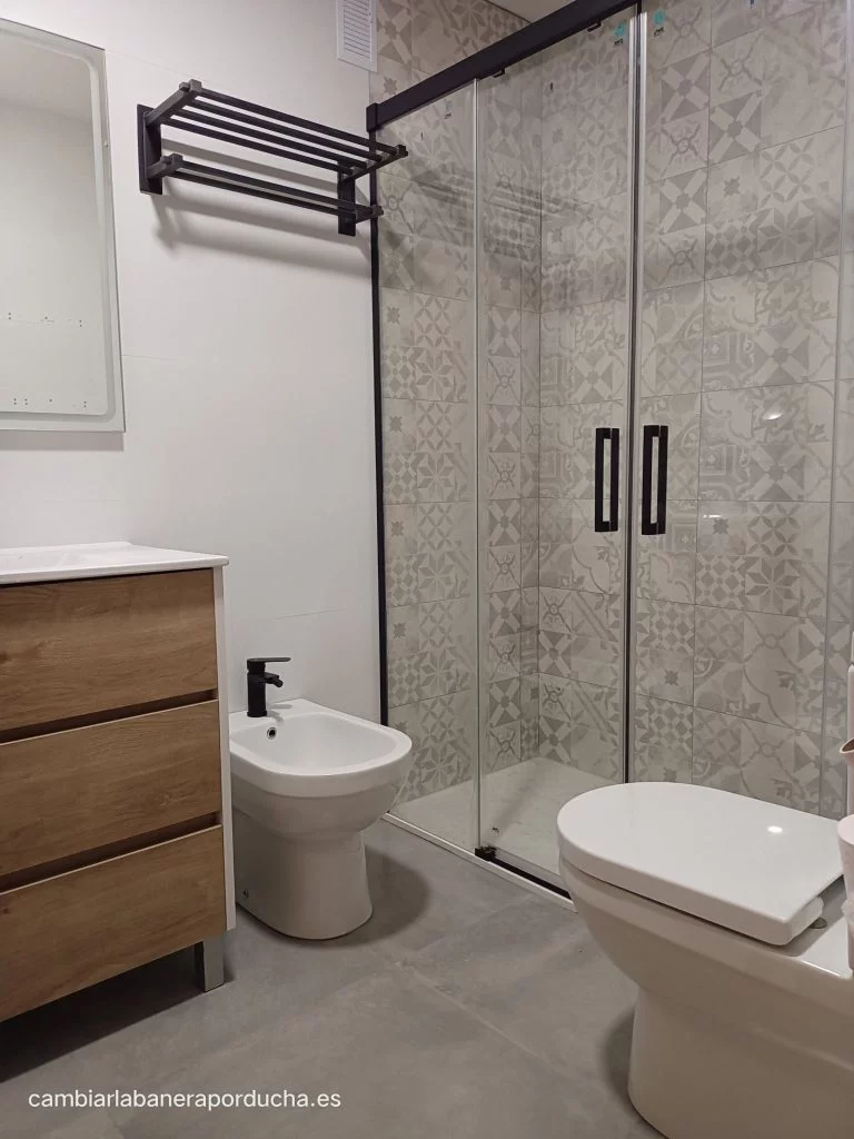 La imagen muestra un baño moderno con una combinación de colores blanco y gris En pamplona. El baño tiene un inodoro, bidé y lavabo blancos con un tocador de madera. La ducha tiene una puerta de vidrio con un mango negro y un patrón de azulejos grises. El baño tiene un toallero y un espejo sobre el lavabo. El suelo está hecho de azulejos grises