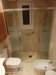 La imagen muestra un baño en el que se ha reemplazado una bañera por una ducha en Logroño. La ducha está hecha de paneles de vidrio con un marco metálico y tiene una puerta corredera. La ducha tiene una ducha y un mango. El baño tiene azulejos beige en las paredes y el suelo. El baño tiene un inodoro y un bidé blancos. El baño tiene un armario de madera con un espejo encima. El baño tiene una pequeña ventana.