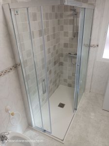La imagen muestra un baño en Galilea (La Rioja) que ha sido remodelado para cambiar la bañera por una ducha. El espacio de la ducha es moderno y elegante, con paredes de azulejos grises claros y blancos que crean un patrón cuadriculado. Hay una puerta de vidrio transparente con bisagras y marcos metálicos que proporciona acceso a la ducha. Dentro del espacio de la ducha, hay una regadera montada en la pared y una manija para controlar el flujo y la temperatura del agua. El piso del área de la ducha es blanco, con un desagüe cuadrado negro visible.