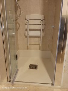 Esta imagen muestra un plato de ducha moderno y renovado en un baño en Orkoien (Navarra), con una silla plegable blanca montada en la pared y una mampara de ducha de vidrio. El plato de ducha es blanco, cuadrado y parece estar hecho de un material liso y duradero. Hay una silla plegable blanca montada en la pared dentro del área de la ducha, probablemente para personas que necesitan sentarse mientras se duchan. Una mampara de vidrio transparente rodea el plato de ducha, con bisagras metálicas brillantes que conectan las piezas individuales del vidrio. Las paredes interiores del área de la ducha están alicatadas con azulejos beige claros que tienen un acabado liso.