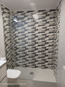 Cambio de bañera por ducha en Pamplona. La imagen muestra un baño moderno con ducha a ras de suelo y mampara de cristal. La ducha tiene un plato de ducha de pizarra natural antideslizante y un grifo termostáyico + kit de ducha. La mampara de ducha es de cristal templado con marco negro y tiene una puerta corredera. El baño está decorado con azulejos blancos y negros