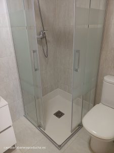La imagen muestra un cambio de plato de ducha por plato de ducha de esquina con puertas de vidrio y una base blanca, que tiene un desagüe cuadrado en el centro. Las paredes están alicatadas con azulejos de color beige claro. A la derecha de la cabina de ducha, se puede ver un inodoro.