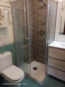 "Reforma de baño en Fuenmayor, La Rioja, mostrando un cambio de plato de ducha por un plato moderno de color claro con mamparas correderas y revestimiento de azulejos en tonos grises y líneas horizontales. La zona de ducha se integra con sanitarios y muebles de baño en madera clara y cerámica blanca."