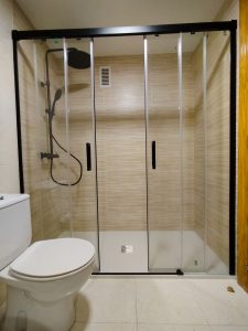 Cambio de bañera por plato de ducha extraplano de 2 cm en Vitoria. Reforma de baño con mampara de cristal corredera con perfilería negra, grifería termostática en acabado negro mate con rociador de lluvia y ducha de mano. Paredes revestidas con azulejos efecto madera en tonos beige. Instalación moderna y funcional para mayor accesibilidad.